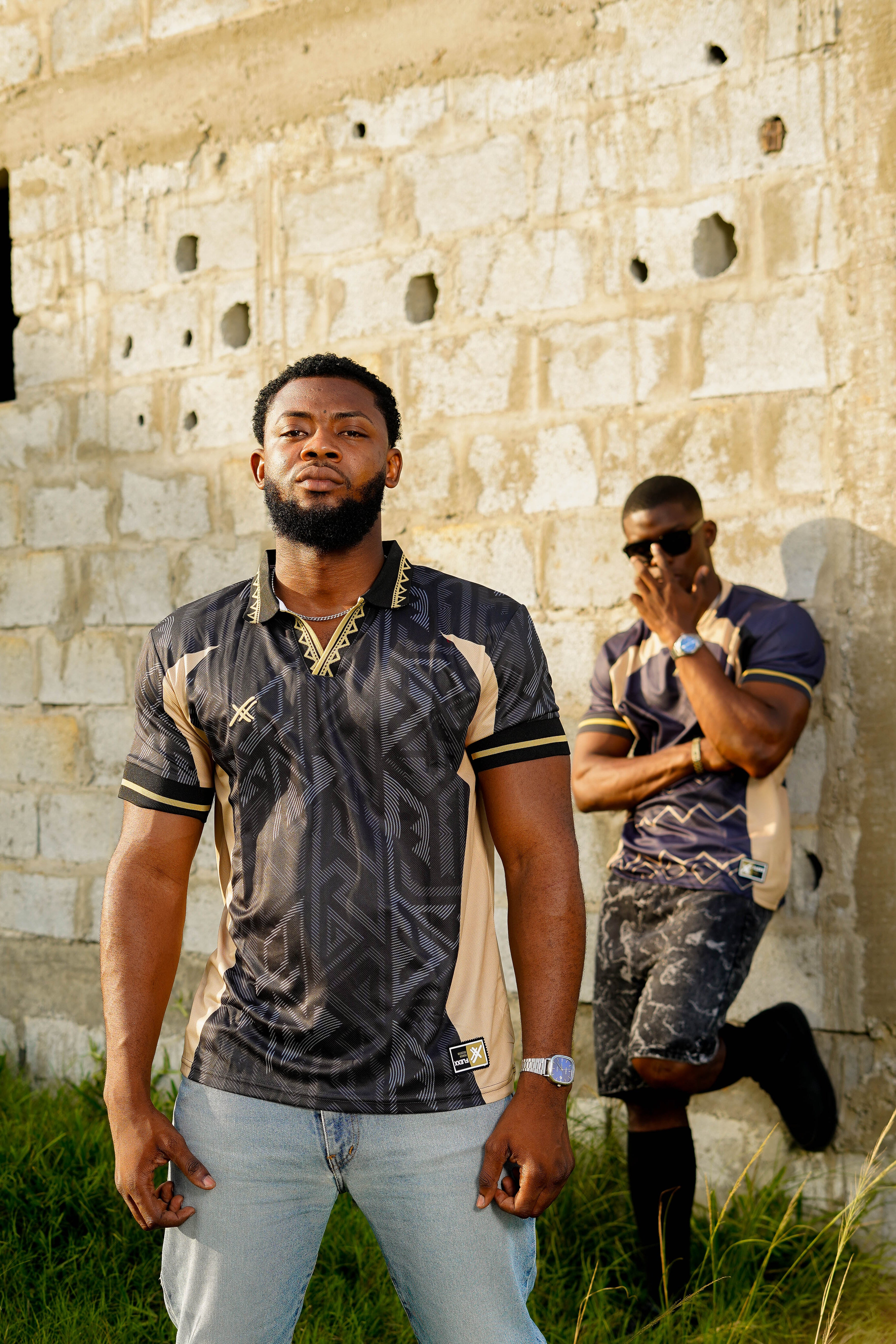 Two men standing in front of a stone wall, one wearing sunglasses and a patterned shirt.